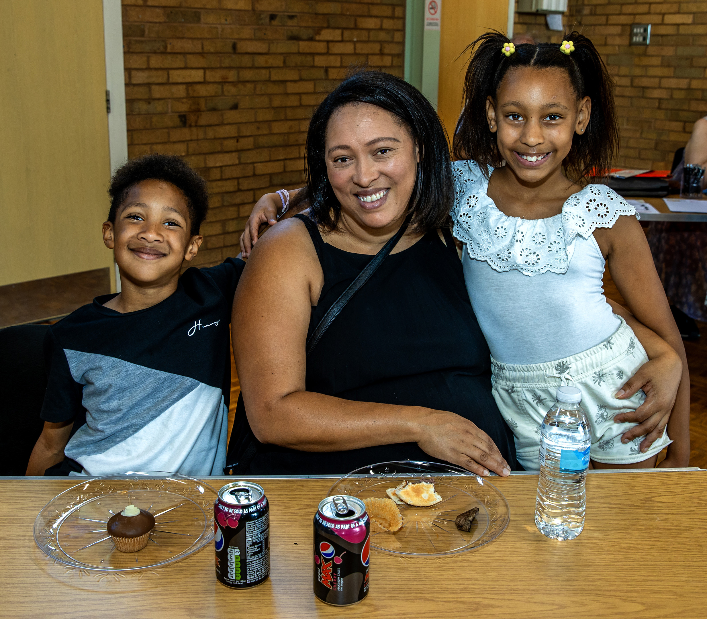 A woman is sat at a table. Her two children, a young boy to her left and young girl to her right, are stood next to her.