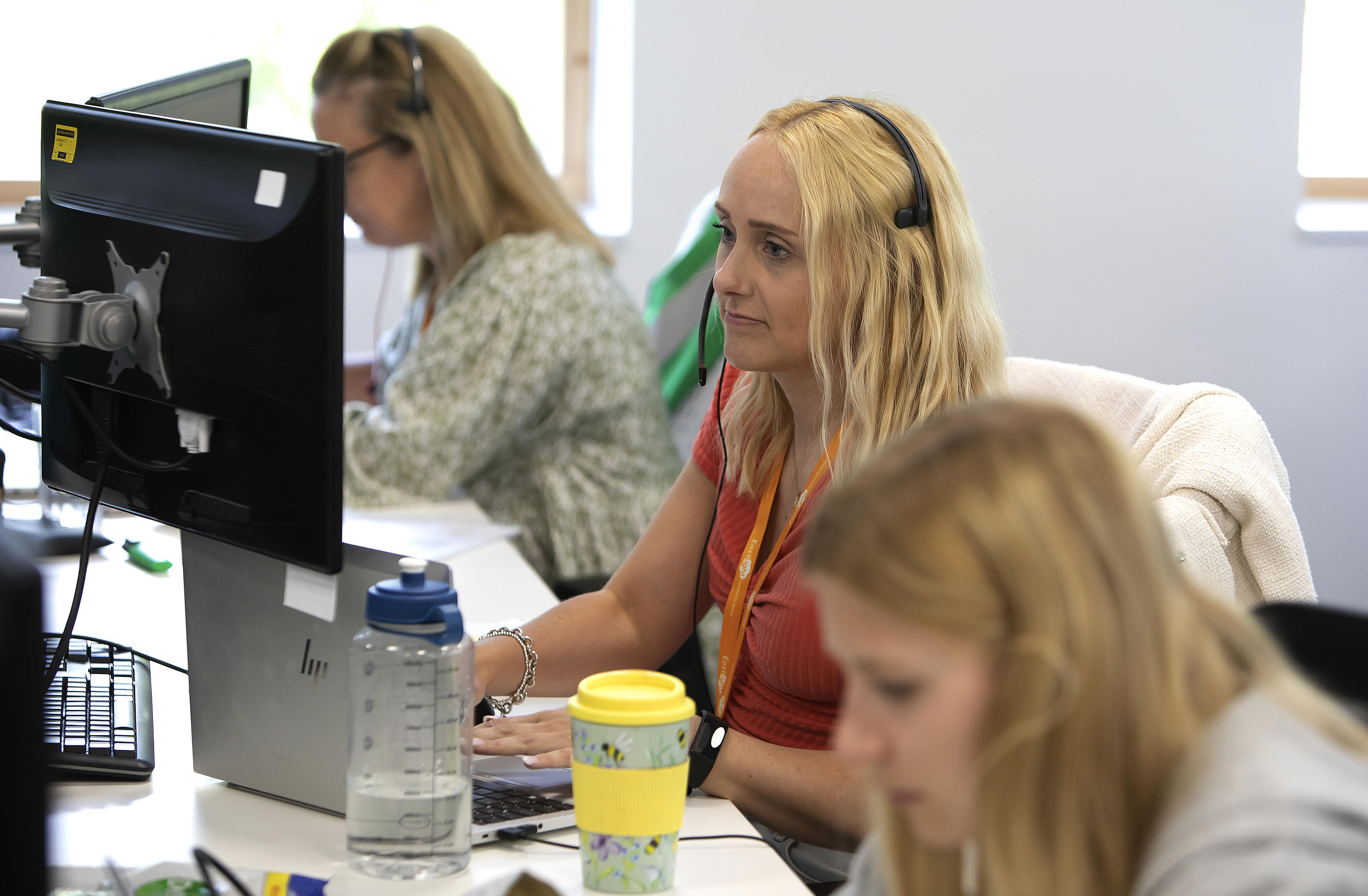 A woman wearing a headset is looking at a laptop screen. There's another woman in the background doing the same.