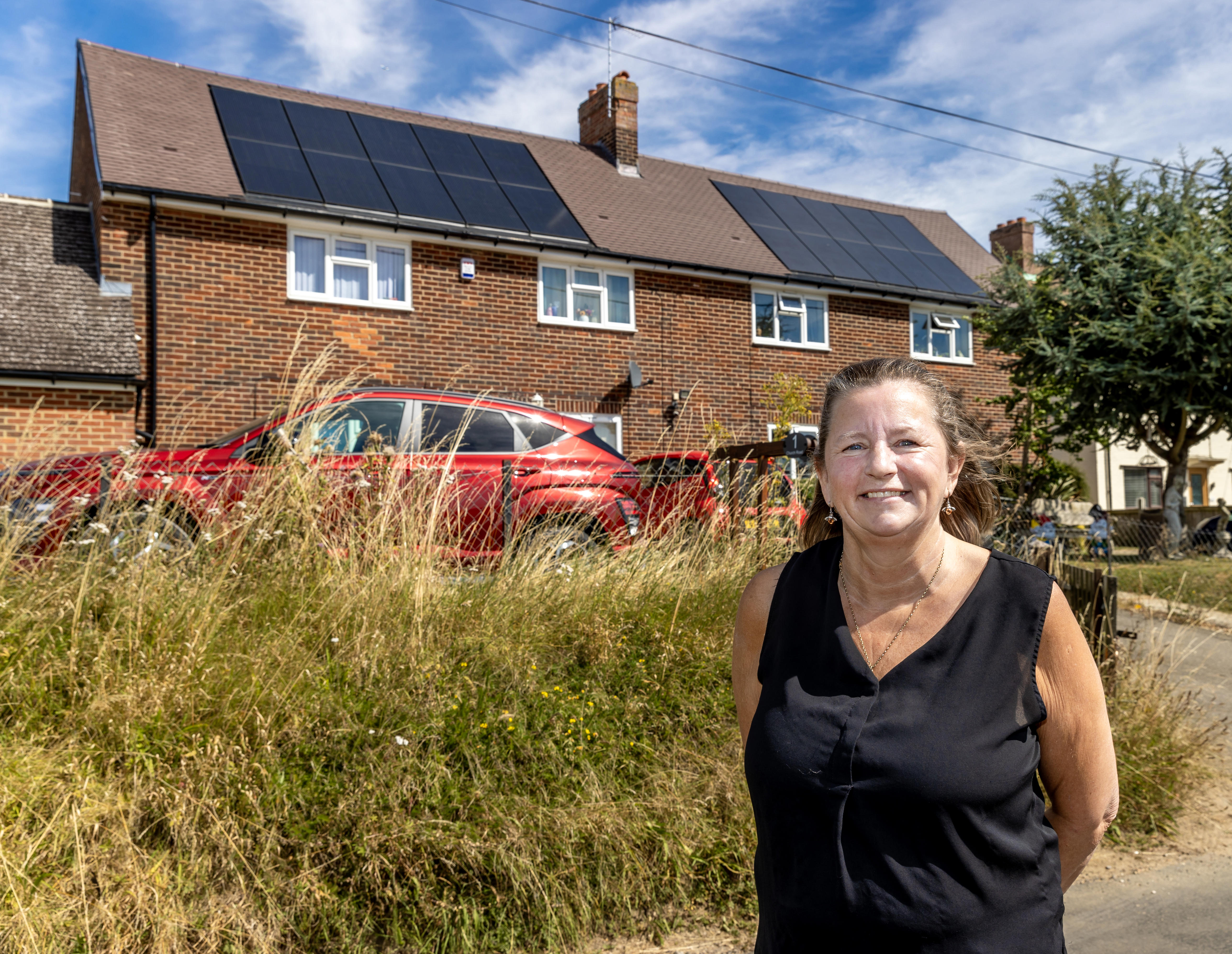 A woman in a black top is standing with her arms behind her back and smiling at the camera. In the background is two houses with solar panels and a red electric car.