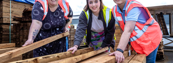 Three women in hi-vis jackets are leaning and picking up timber board.