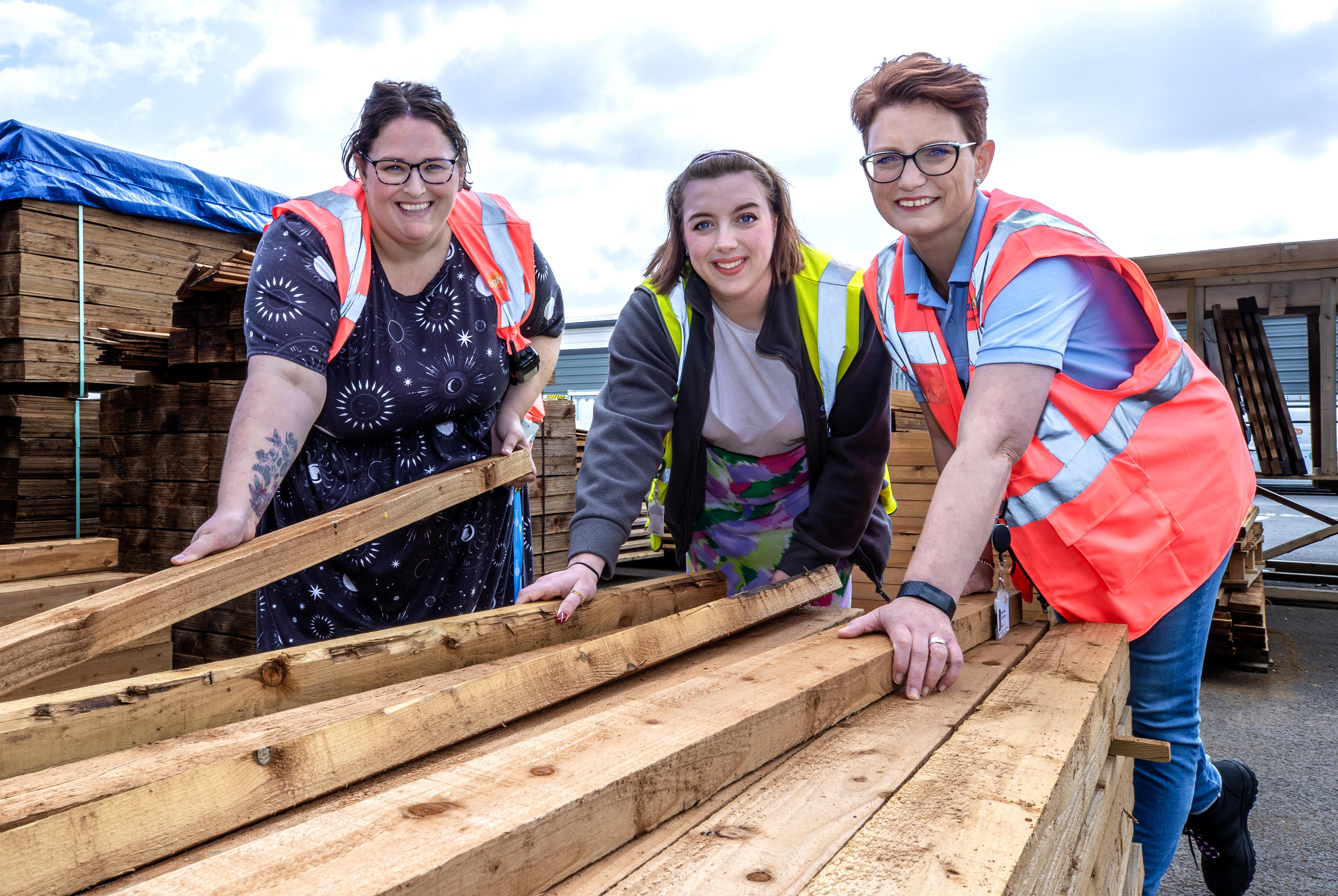 Three women in hi-vis jackets are leaning and picking up timber board.