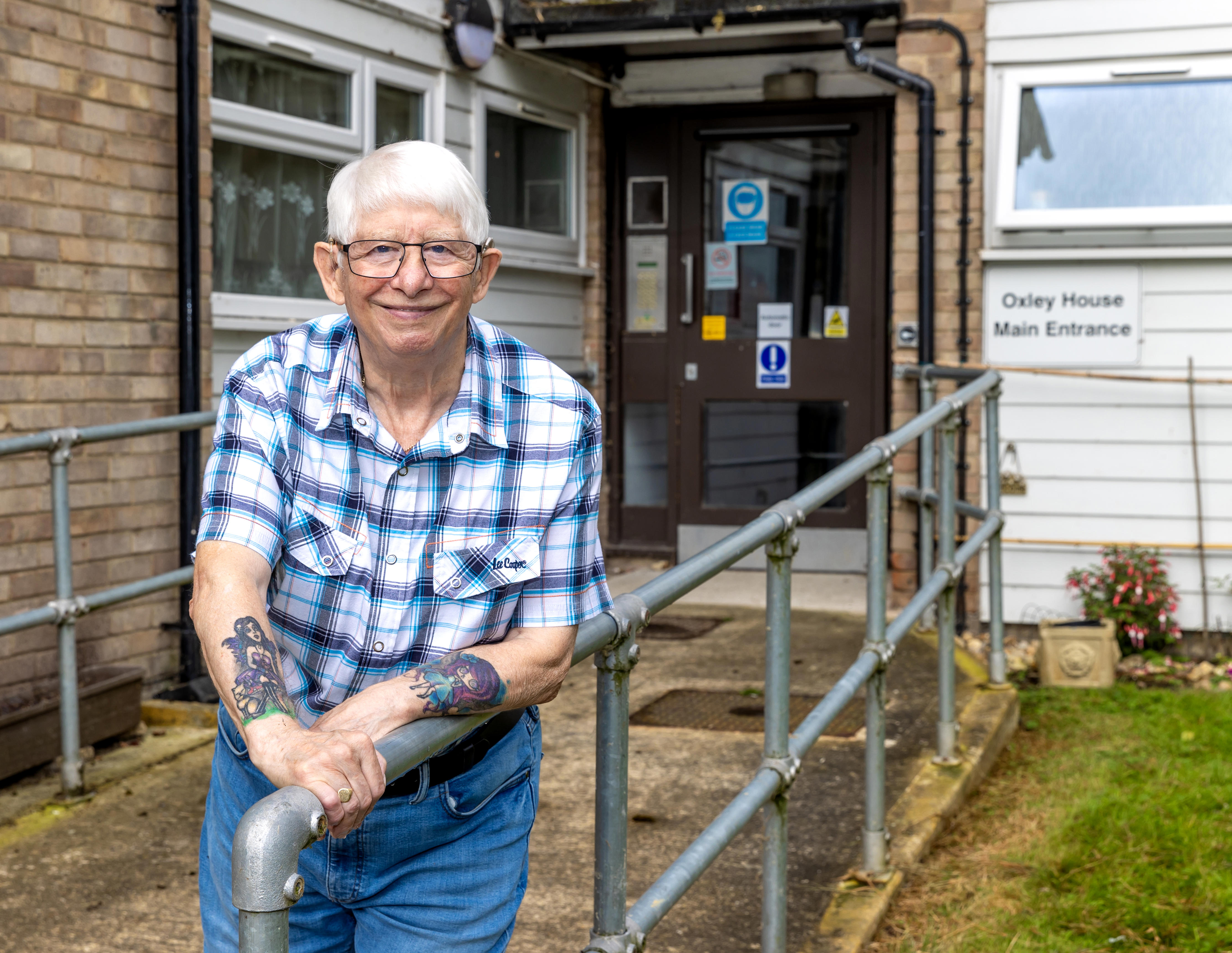 An older man with glasses is leaning on a metal railing which leads up a ramp to a block of flats.