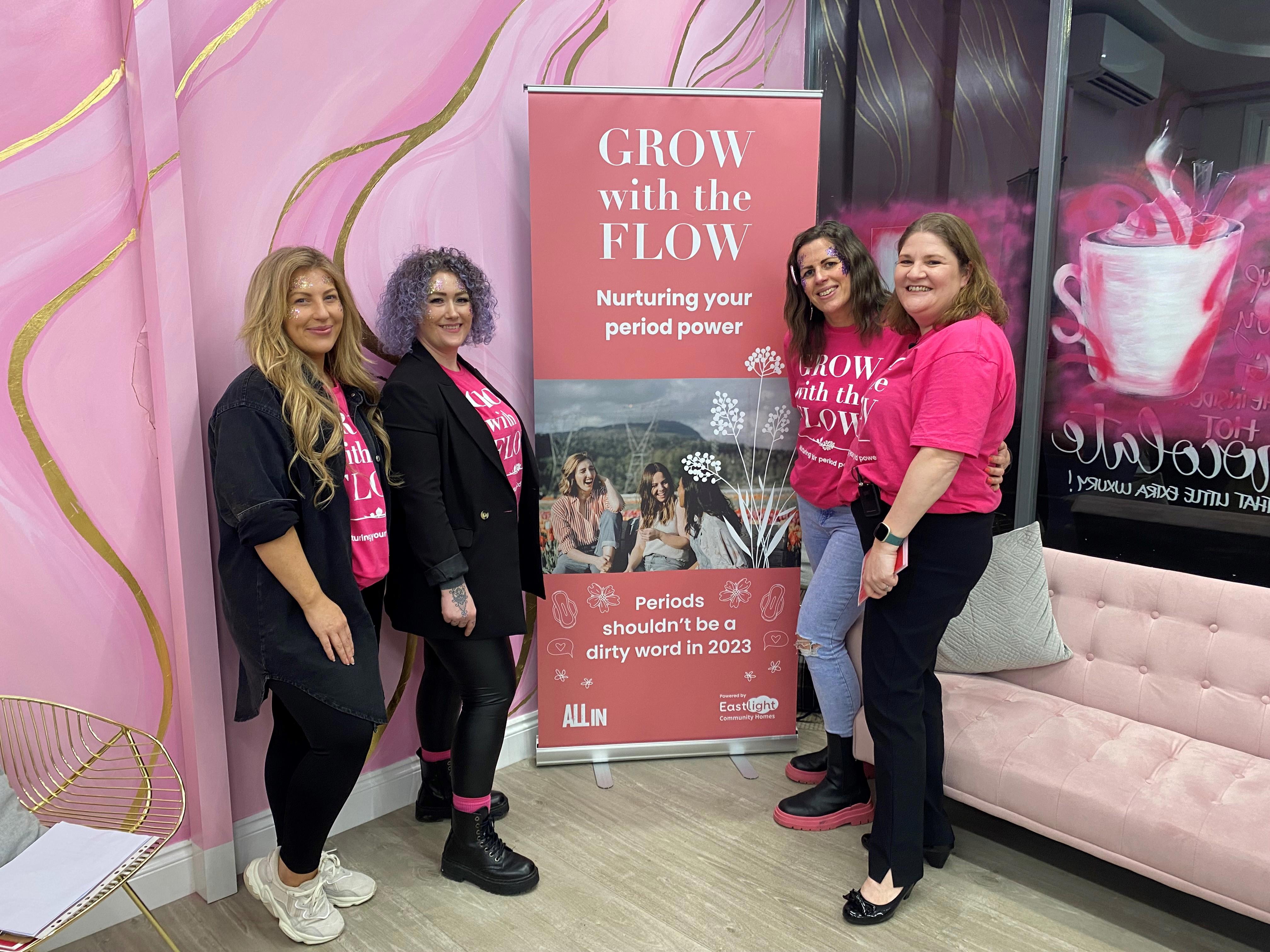 Four women are stood around a banner. They are each wearing pink tshirts that says 'Grow With The Flow'