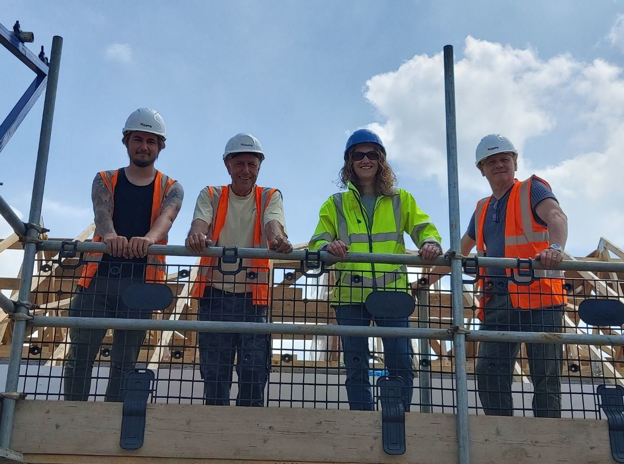 Eastlight Community Homes Development Manager with partners on a building site. Scaffolding is erected, and four people in hard hats and hi vis look down at the camera holding onto a railing and smiling