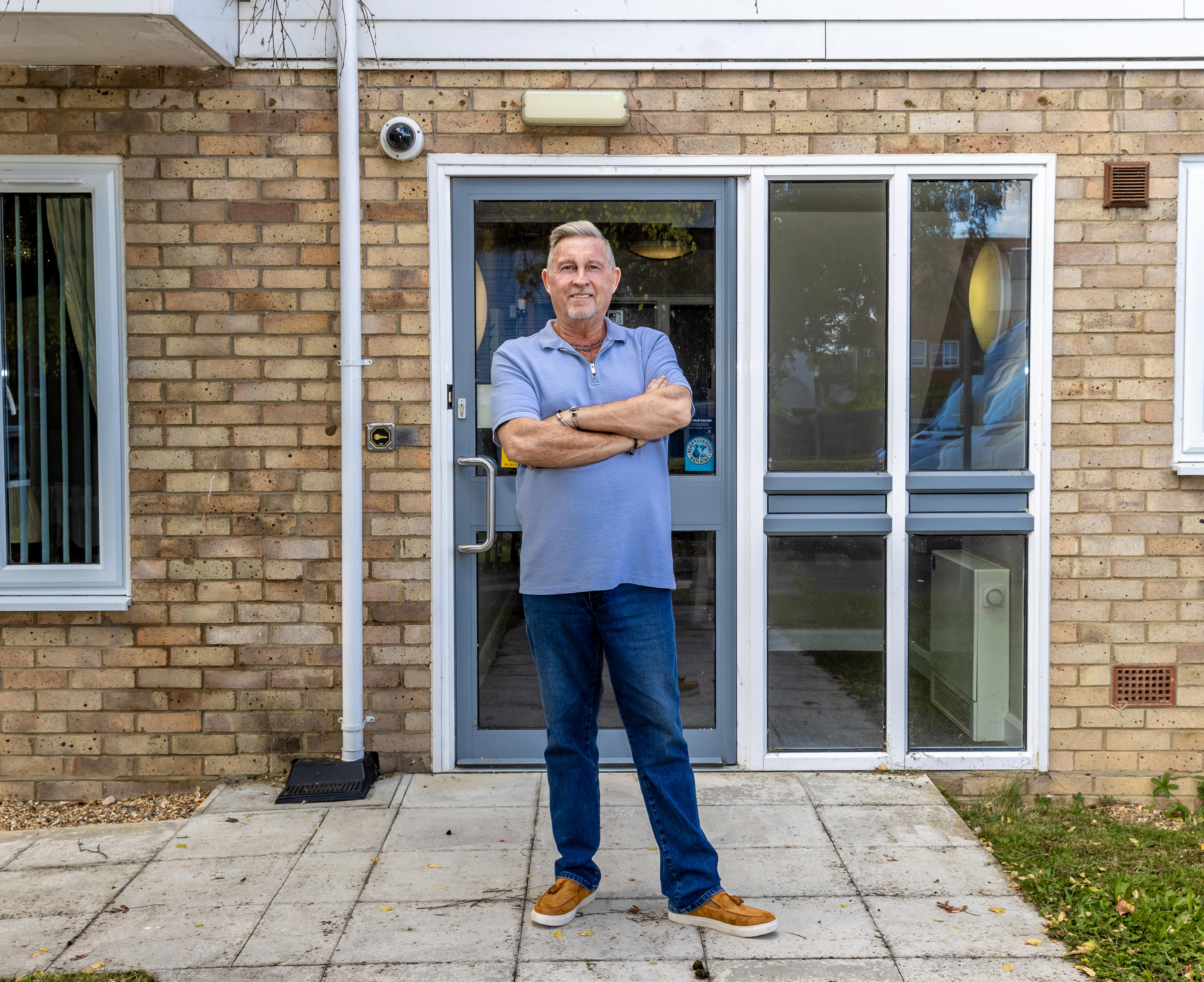 A man is stood with his arms folded in front of a door to flats.