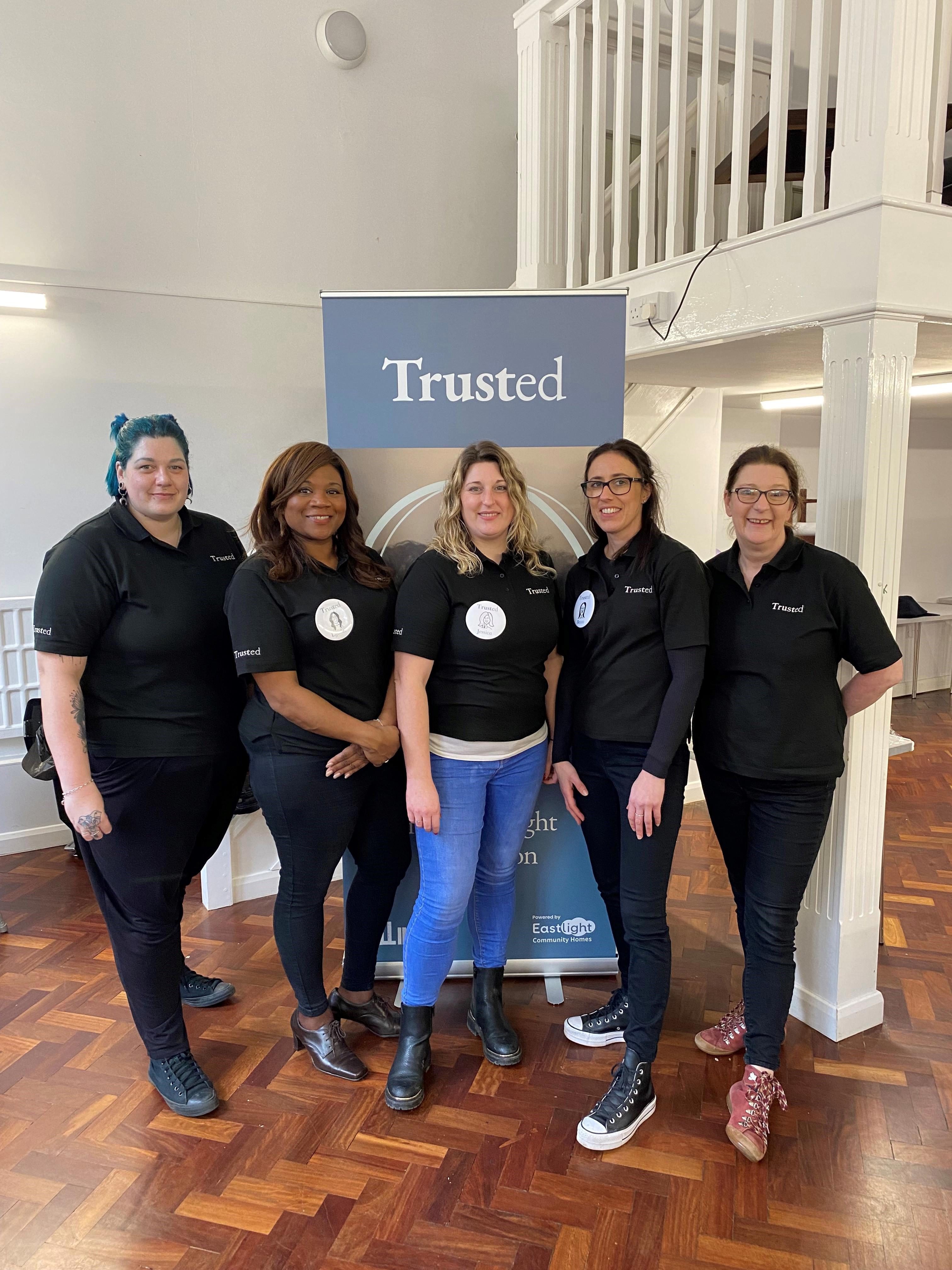 Five women are stood in a line. They are all wearing black polos with 'Trusted' embroidered on them.