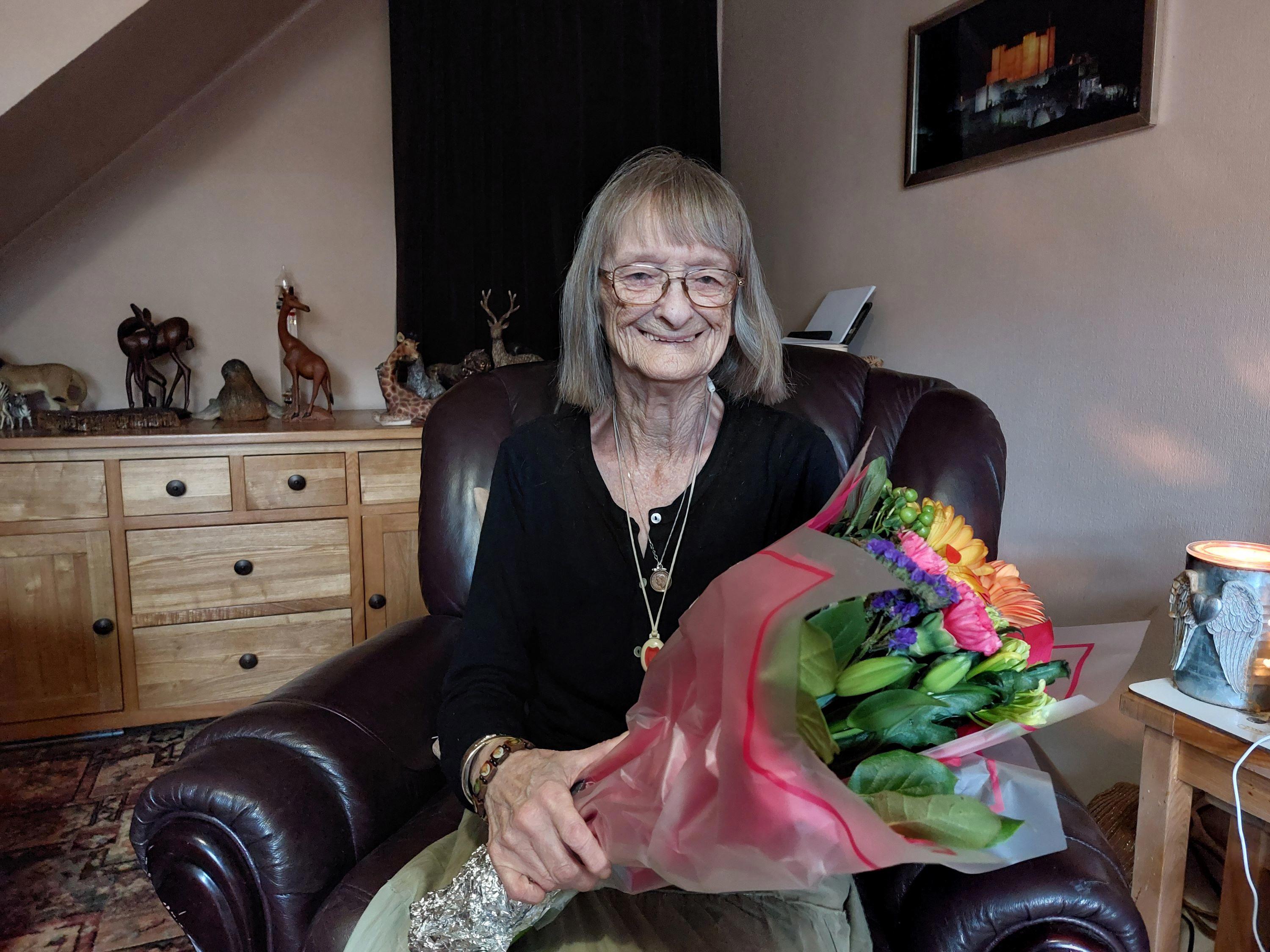 An older woman is sat in an armchair in her home. She is holding a bouquet of flowers.