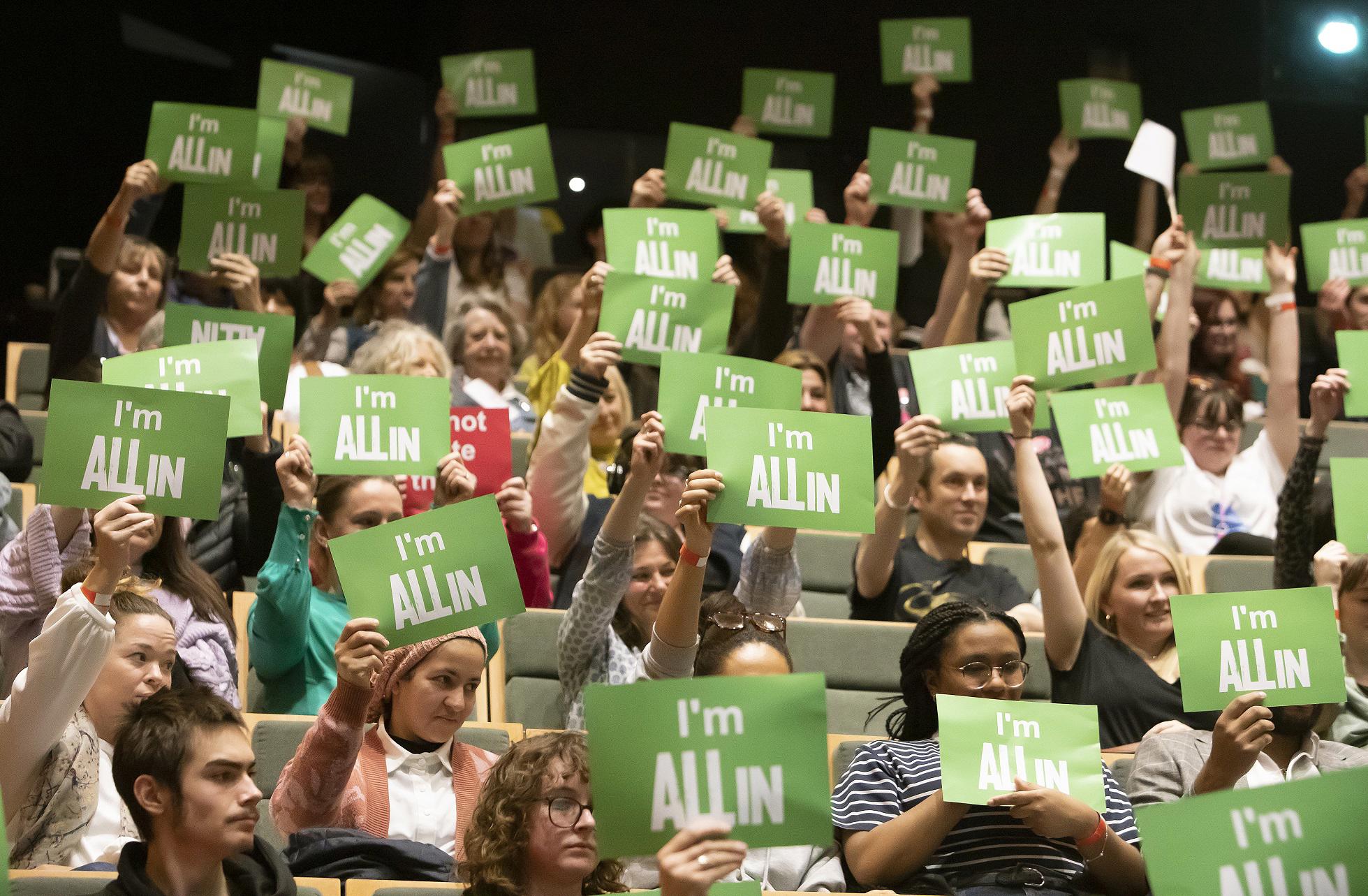 An audience of people are holding up pieces of green paper which say "I'm All In"