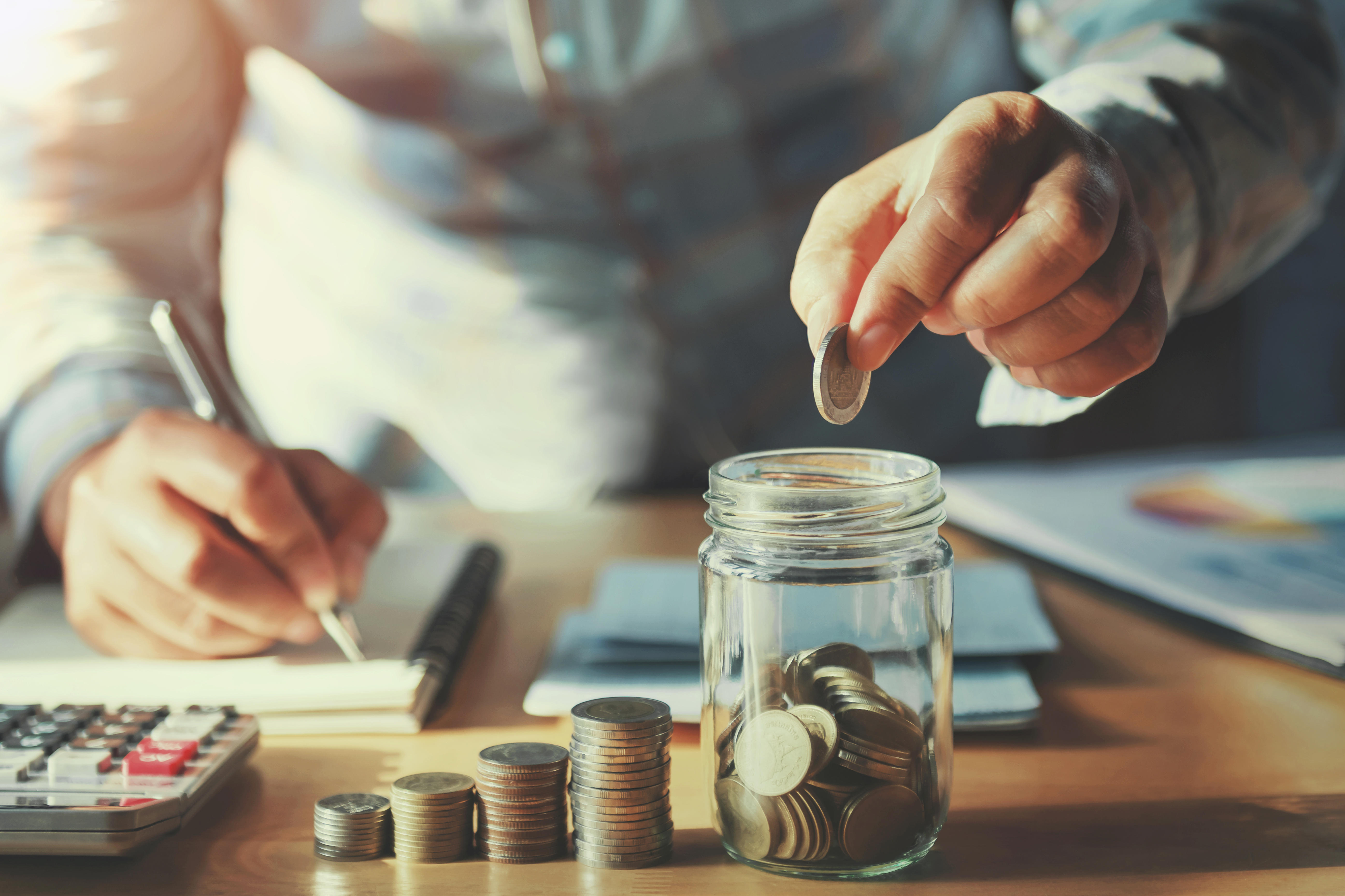 A glass jar with coins in is being add to by a hand. There are stacked coins next to the jar.