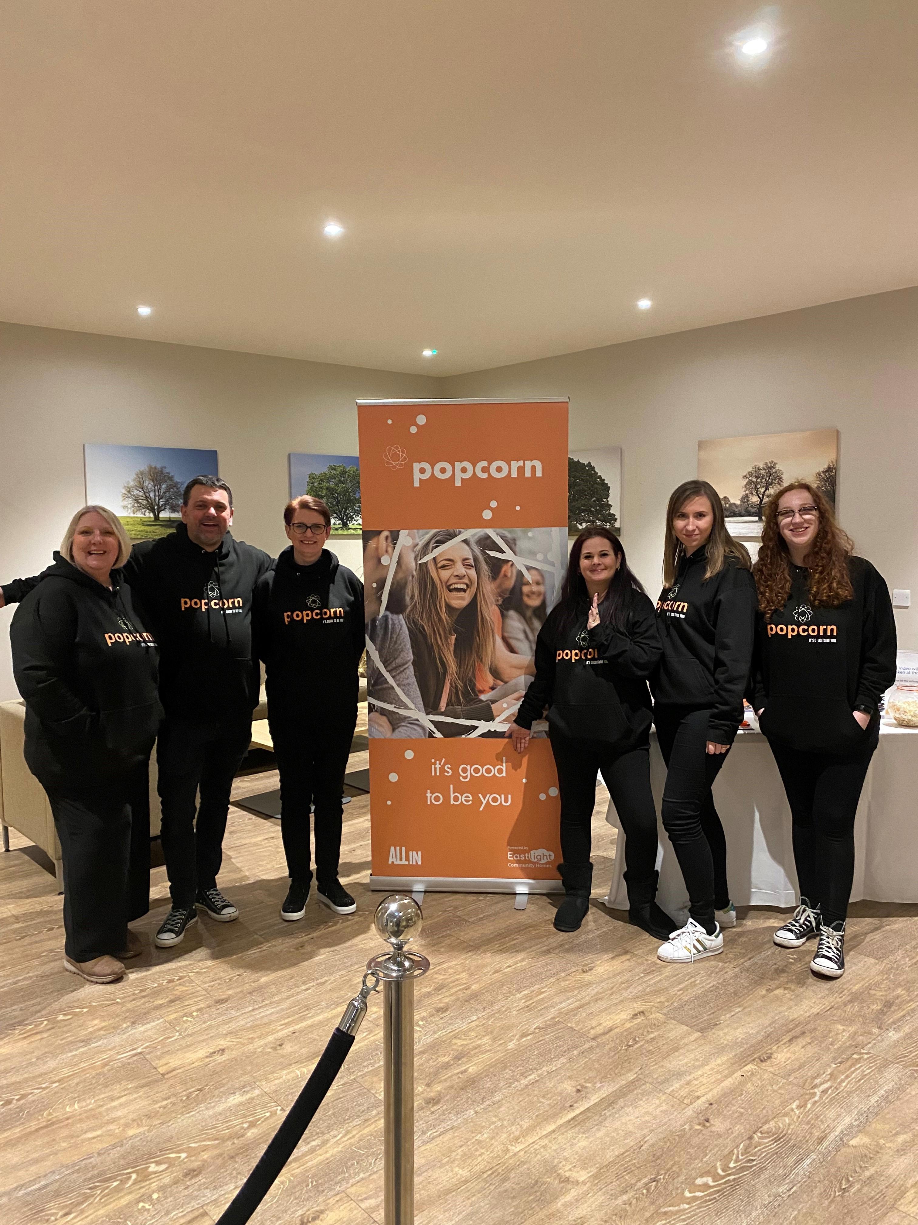 5 women and 1 man are stood in a line next to a banner. They are all smiling at the camera and wearing black hoodie with the word 'Popcorn' printed on the front.