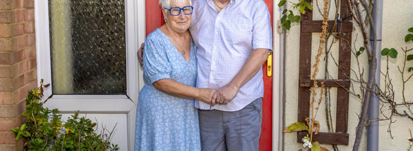 An older couple are stood on their door step holding hands and smiling at the camera.