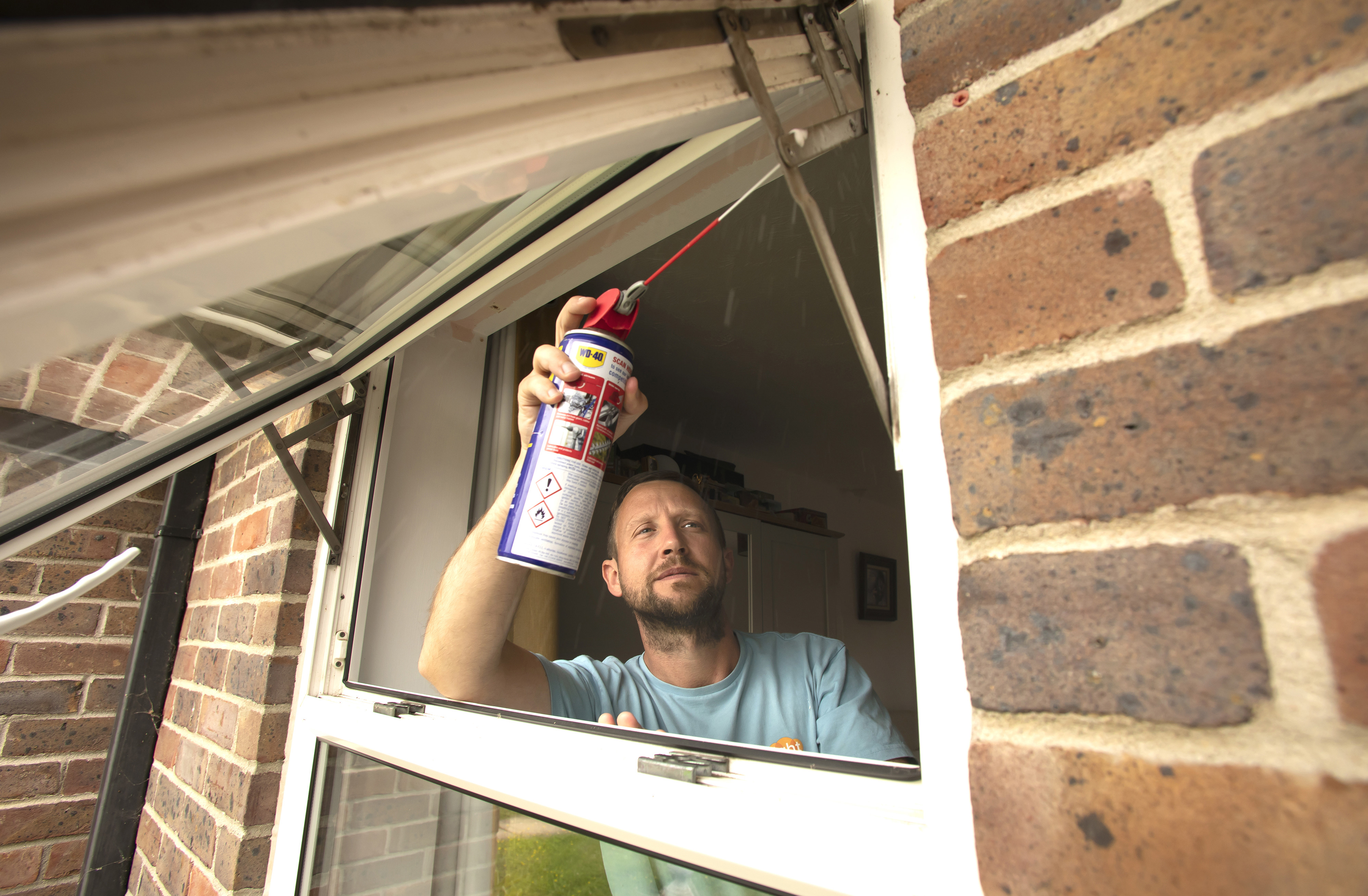 A man wearing an Eastlight tshirt is spraying a lubricant onto a window hinge.