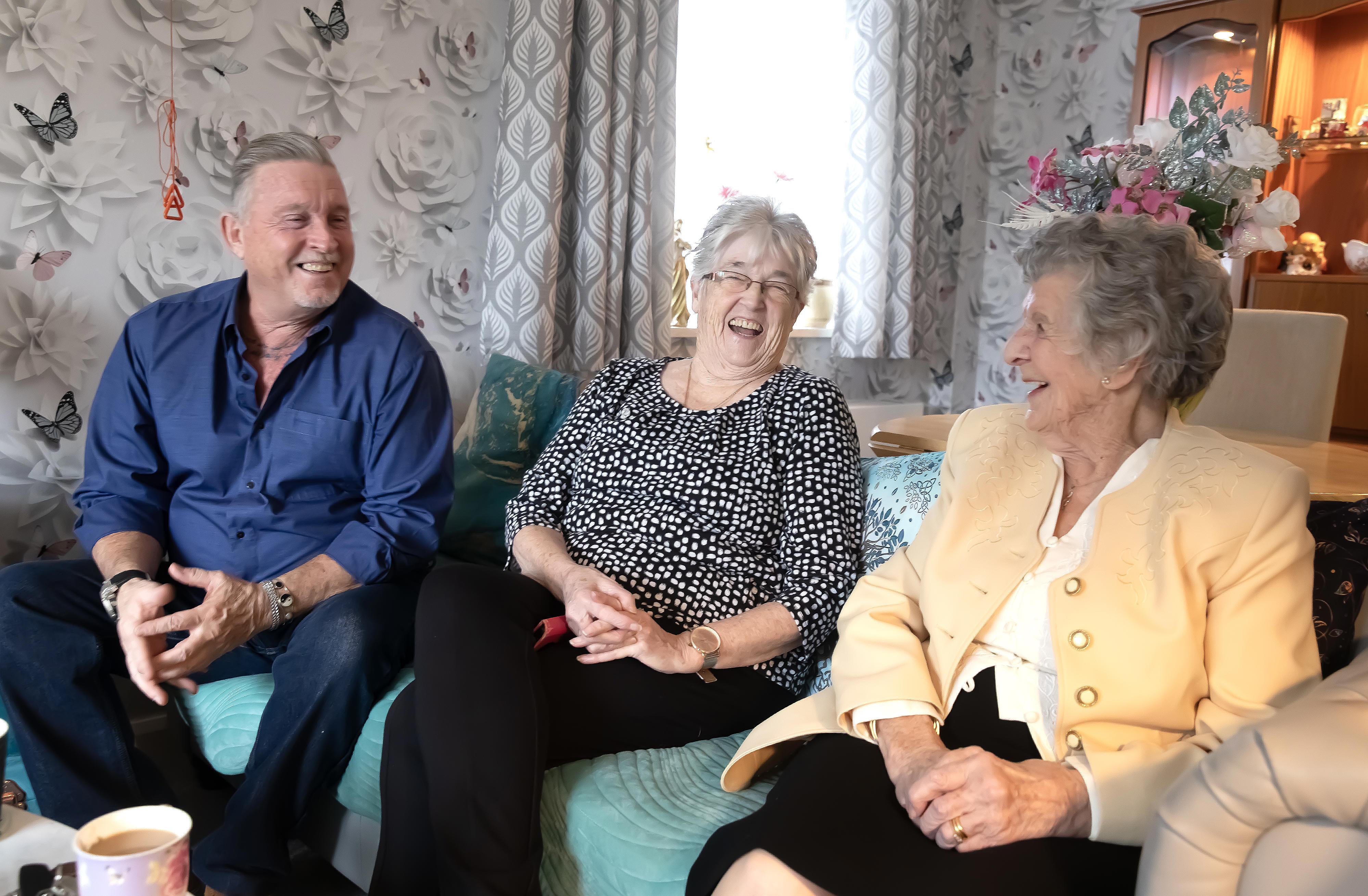 An older man and two older women are sat on a sofa laughing.