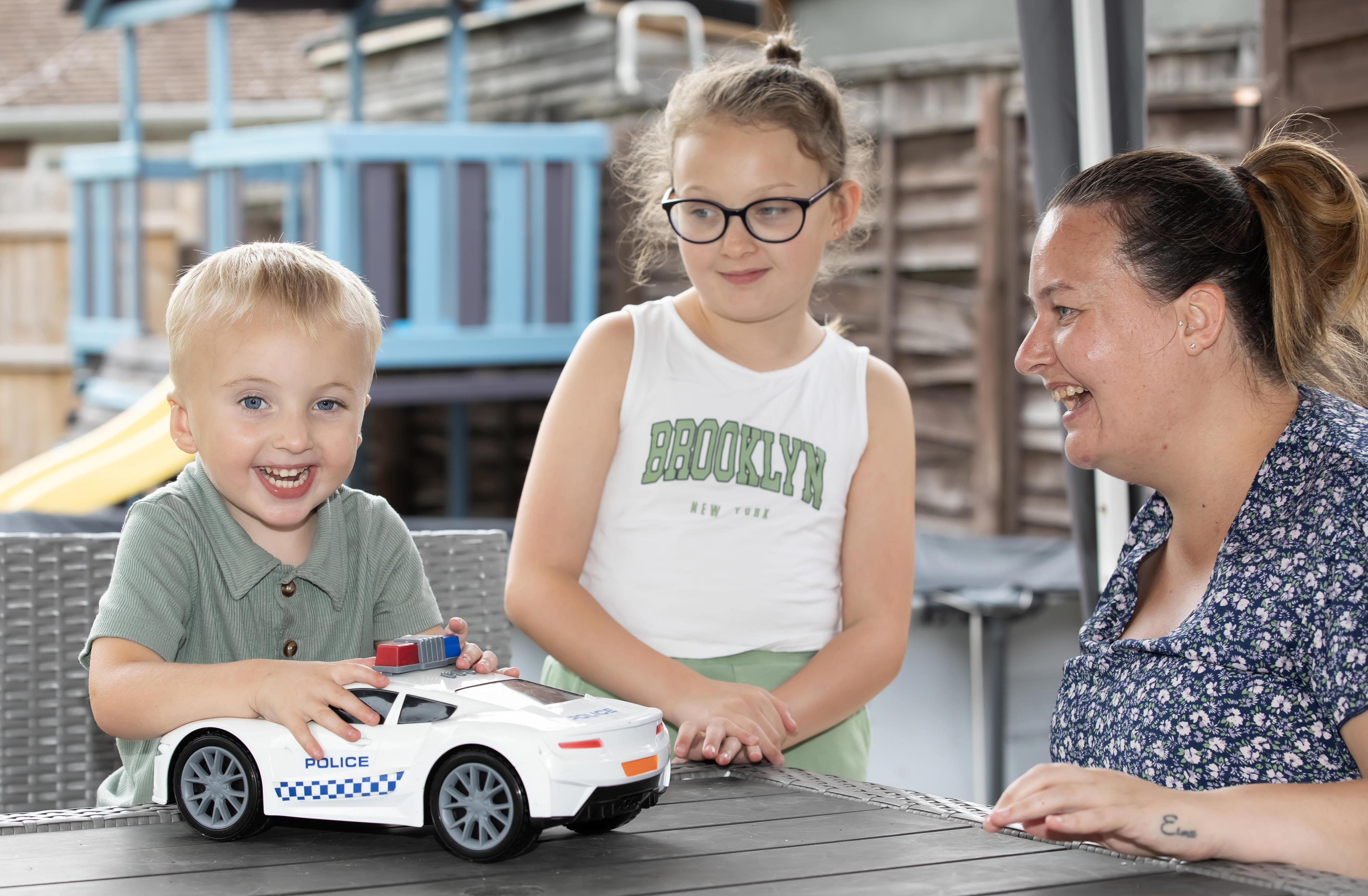 A woman with her two young children are playing on a table. The young boy is holding a large police car toy.